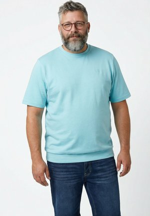 Mężczyzna w średnim wieku z siwą brodą i okularami, ubrany w jasnoniebieską koszulę z krótkim rękawem i ciemnoniebieskie dżinsy, stojący na jednolitym tle.