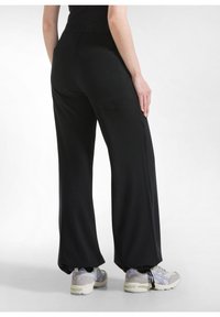 Wijdvallende zwarte broek met een gladde textuur, een elastische tailleband en twee achterzakken, gecombineerd met lichtgekleurde sportschoenen.