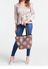 Brązowa torba tote z białymi wzorami geometrycznymi, z czarnymi uchwytami i eleganckim wzornictwem. W połączeniu z bluzką w kropki i dżinsami.