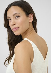 Vest top in light cream fabric with a V-neckline and decorative knot detail on the straps. Smooth texture enhances the overall design.