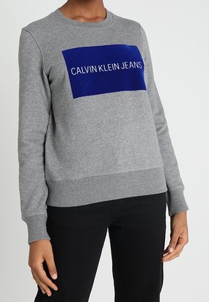 Persoon draagt een grijze trui met een blauw vierkant en de tekst "CALVIN KLEIN JEANS", gecombineerd met een zwarte broek, staand tegen een effen achtergrond.