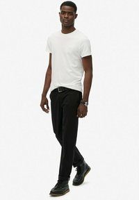 Camiseta blanca de manga corta, pantalones negros y zapatos oscuros con superficie texturizada. Conjunto informal con un ajuste elegante y diseño minimalista.