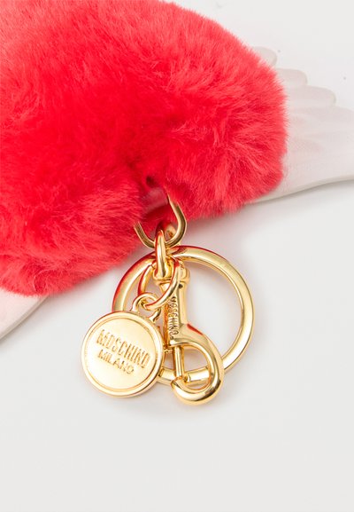 Porte-clés rouge duveteux avec anneaux dorés et une étiquette ronde en or gravée de "MOSCHINO MILANO" sur une surface blanche.