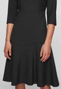 Svart klänning med figurnära liv, trekvartslånga ärmar och utsvängd nederkant. Tillverkad av slät, stretchig tyg med en enfärgad design.