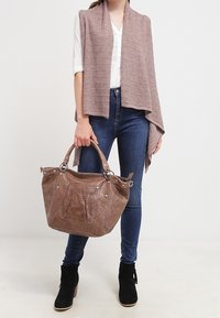 Braune Handtasche im Krokodilmuster mit stabilen Griffen, kombiniert mit einem hellrosa Wickel und dunklen Jeans, vor einem neutralen Hintergrund.