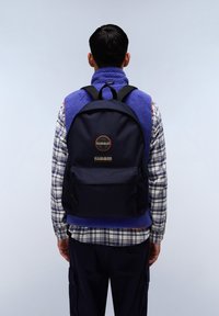 Marinblått tyg ryggsäck med svarta remmar, framficka och logodetalj. Modellen bär en lila fleeceväst över en rutigt skjorta.