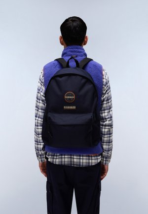 Zaino di tessuto blu navy con spallacci neri, tasca frontale e dettaglio con logo. Il modello indossa un gilet in pile viola sopra una camicia a quadri.