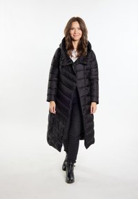 Sort puffers jas med et langt, quiltet design, stor hætte og frontlynlås. Båret over en hvid bluse og sorte bukser, parret med sorte ankelstøvler.