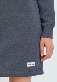 Robe sweatshirt grise en matériau doux, dotée de manches longues et d'une étiquette blanche distinctive avec "oxmo" au bas de l'ourlet.