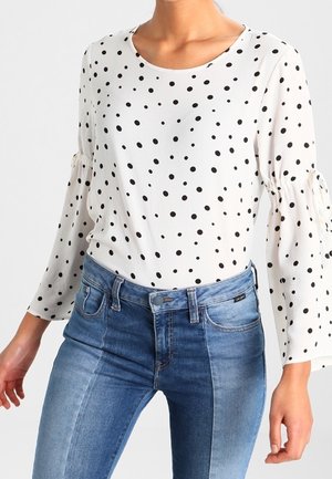 Maglietta bianca a maniche lunghe con pois neri, scollatura rotonda e polsini arricciati, abbinata a jeans in denim blu chiaro.
