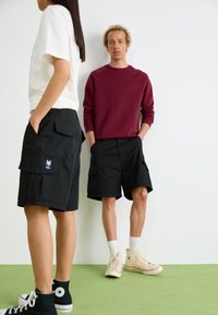 Sorte cargo shorts med sidelommer, båret af en person i en hvid t-shirt og en anden i en vinrød sweatshirt. Lys grøn gulv.
