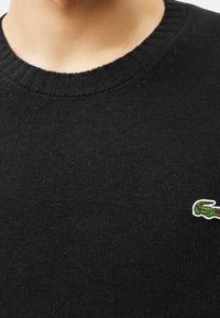 Svart stickad tröja med ribbat krage och en liten grön och vit krokodil-logotyp på vänster sida. Mjuk textur, avslappnad design.