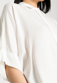 Camicia bianca leggera con colletto e chiusura a bottoni. Presenta maniche corte arrotolate e una leggera trama su tutto il tessuto.