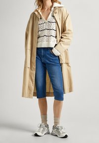 Světlá béžová dlouhá pláštěnka přes bílý pletený svetr s námořnickými vzory, doplněná modrými capri džíny a šedými teniskami s bílými detaily.