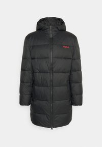 Schwarze gesteppte Jacke mit Reißverschluss vorne, Kapuze und rotem "HUGO"-Logo. Verfügt über eine kastige Form und eine glatte Textur.