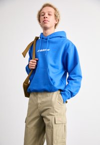 Blå hoodie med framficka och vit logotyp, matchad med beige cargobyxor med sidfickor och sömdekor.