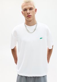 Witte T-shirt met korte mouwen van een ademende stof, met een subtiele groene logo, ronde halslijn en een relaxte pasvorm, gecombineerd met grijze broek.