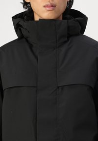 Black waterproof jacket with a high collar, featuring a seamless design and minimal front detailing for a sleek appearance.