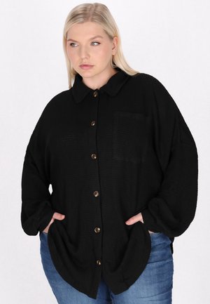 Camicia nera con bottoni, colletto, tessuto strutturato e taschino sul petto. Presenta bottoni marroni e una vestibilità comoda. Abbinata a jeans blu.