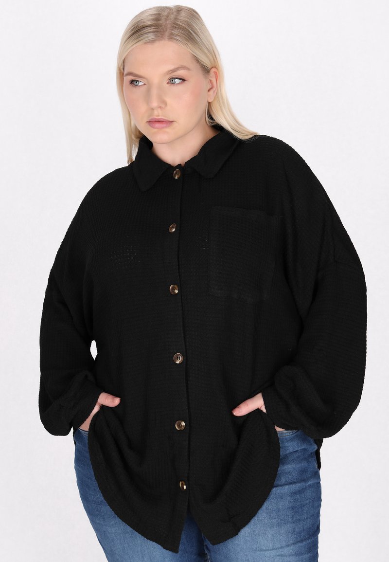 Camicia nera con bottoni, colletto, tessuto strutturato e taschino sul petto. Presenta bottoni marroni e una vestibilità comoda. Abbinata a jeans blu.