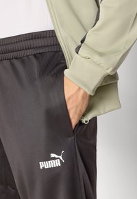 Puma træningsdragt med en olivengrøn zip-jakke og sorte bukser, lavet i et struktureret stof og med hvidt Puma-logo på venstre ben.
