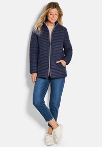 Marineblauwe gewatteerde jas met een hoge kraag, voorrits en twee zijzakken. Gecombineerd met blauwe jeans en witte sneakers.