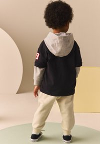 Enfant portant un t-shirt noir à manches courtes avec une capuche grise, un pantalon de survêtement crème et des baskets noires avec des semelles blanches. Détail en numéro rouge sur le t-shirt.