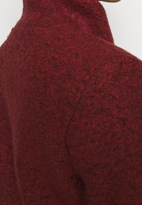 Top de mezcla de lana rojo de cuello alto con una superficie texturizada, que presenta un diseño ajustado que resalta la zona del cuello y los hombros.