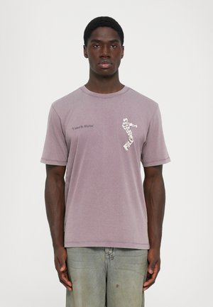 Man wearing faded purple T-shirt with "United By Rhythm" text and trumpet player graphic, paired with worn light gray jeans.