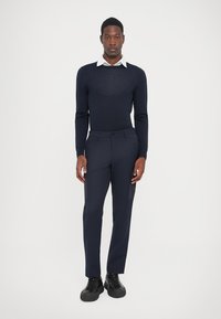 Bărbat tânăr în picioare, îmbrăcat cu pantaloni slim-fit bleumarin, pulover bleumarin peste o cămașă albă cu guler și adidași negri, pe un fundal alb simplu.