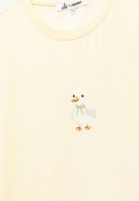 Camiseta de algodón amarillo claro que presenta un pequeño pato bordado con un lazo verde, diseñada con cuello redondo y mangas cortas.