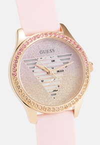 Rosa silikonremme med en rund glitrende sølvskive, guldfarvet bezel og rhinestone-dekorationer omkring et trekantet mønster.