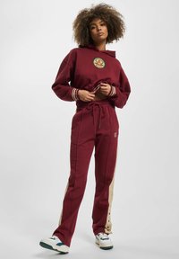 Bordeaux hættebluse med logo og stribede manchetter, parret med matchende bordeaux joggers med beige sidepaneler. Hvide sneakers med grønne accenter.
