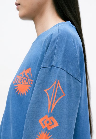 Modrá oversized mikina má na prednej strane a na rukávoch oranžový grafický text a geometrické vzory. Materiál má mäkký, textúrovaný vzhľad.