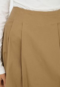 Jupe plissée kaki en coton, dotée d'une taille haute et d'une texture lisse, avec des poches latérales et une fermeture éclair dissimulée.