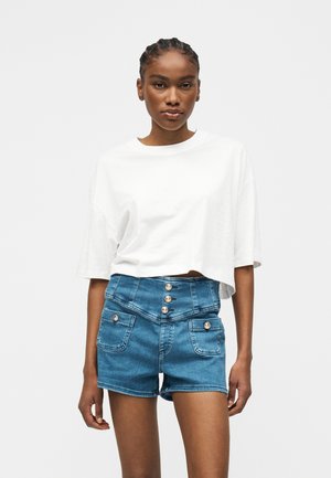 Młoda kobieta z warkoczami, ubrana w luźny biały krótki top i niebieskie szorty z denimu z wysokim stanem, z guzikami i przednimi kieszeniami.