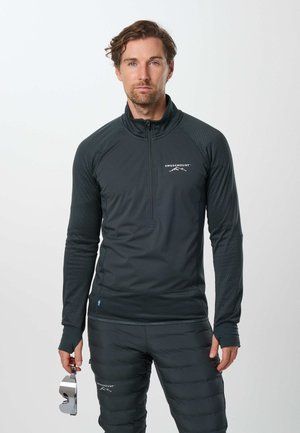 Homme en pantalon matelassé noir et haut zippé noir à manches longues tenant des lunettes de ski argentées sur un fond clair uni.