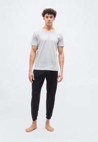 Νεαρός άνδρας με σγουρά μαλλιά στέκεται ξυπόλητος φορώντας ένα ανοιχτό γκρι κοντομάνικο πουκάμισο henley και μαύρο παντελόνι jogger μπροστά από λευκό φόντο.