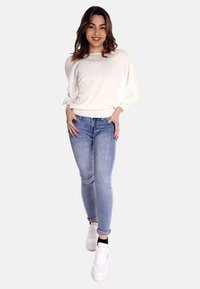 Maglione bianco lavorato a maglia con vestibilità larga, abbinato a jeans skinny blu chiaro e sneakers bianche. Modella in piedi con uno sfondo neutro.