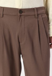 Brown pleated trousers featuring side pockets and a button closure, made of textured fabric with a clean, tailored silhouette.