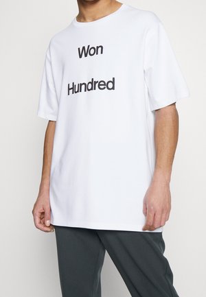 Osoba ubrana w białą koszulkę z czarnym napisem "Won Hundred" w połączeniu z ciemnymi spodniami na jednolitym tle.