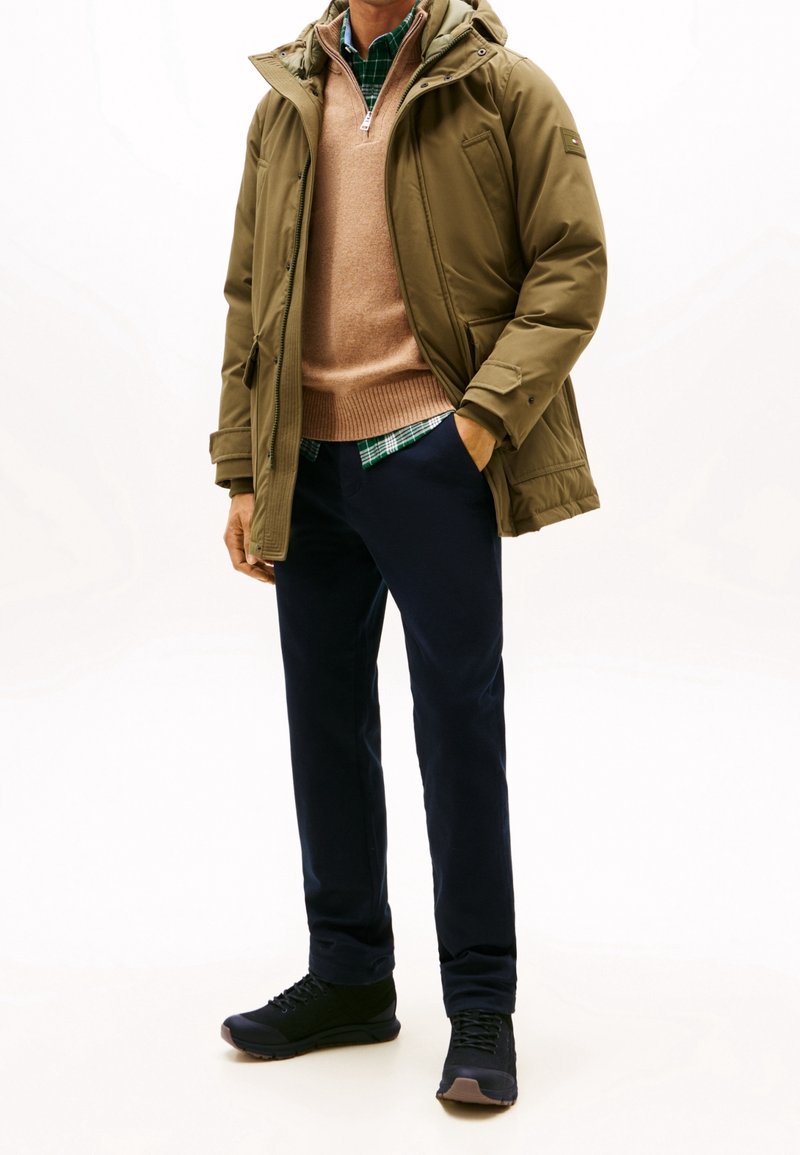 Homme portant une veste à capuche vert olive, un pull beige, une chemise à carreaux verts, un pantalon bleu foncé et des baskets noires, debout avec une main dans la poche.