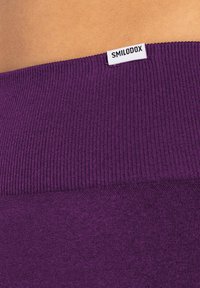 Lila gerippter Stoff mit glatter Textur, ausgestattet mit einem weißen Etikett, auf dem "SMILODOX" am oberen Rand steht. Nahaufnahme des Bunds.