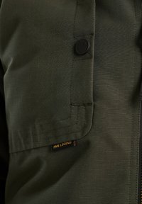 Groene stoffen jas met een gestructureerd oppervlak, een opvallende zwarte knop en een zichtbaar PME Legend label met gele letters.