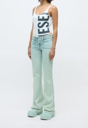 Femme portant un haut blanc sans manches avec un grand texte noir, un jean évasé bleu clair et des chaussures épaisses bleu clair assorties, debout devant un fond blanc.