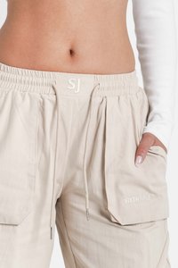 Beige cargobroeken hebben een elastische tailleband met trekkoord, zijzakken en geborduurde logo-details aan de voorkant. Gestructureerde stof.
