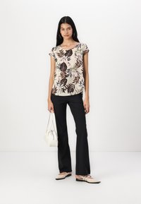 Top à imprimé floral en crème et marron avec des manches courtes, associé à un pantalon évasé noir, des ballerines beiges et un sac à main blanc. Fond simple.