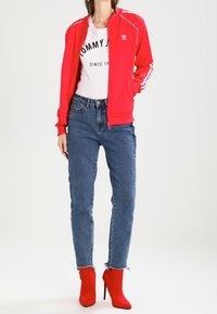 Červená sportovní bunda s bílými pruhy, bílé tričko s grafikou, džíny s vysokým pasem a červené kotníkové boty na vysokém podpatku.