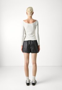 Vit off-shoulder, figurnära långärmad topp i kombination med svarta shorts i konstläder. Modellen har på sig vita strumpor och svarta sneakers. Minimalistisk bakgrund.