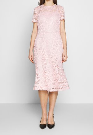 Kobieta w różowej koronkowej sukience do kolan z krótkim rękawem i kwiatowym wzorem oraz czarnych szpilkach z ostrym noskiem, stojąca na jednolitym tle.